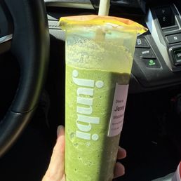 Blended Matcha - A person holds a large green matcha drink in a clear plastic cup with a straw, inside a vehicle. The cup has the brand name 'Junni' printed on it and a label indicating a dine-in order for Jenny. The vehicle's dashboard and steering wheel are visible in the background, along with various controls and buttons.
