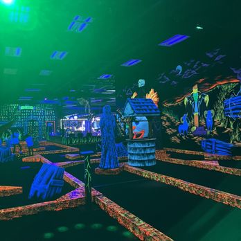 Monster Mini Golf - A blacklight mini golf course features illuminated pathways, animated monster decorations, and vibrant murals on the walls, with a bar area visible in the background. - Mini Golf Near Me - Lafayette, Indiana