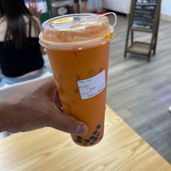 Starry Moon - Nice - A hand is holding a large, clear plastic cup filled with Thai tea, which has a bright orange color and contains black tapioca pearls at the bottom. The cup has a plastic lid and a straw. The background shows a cafe setting with a person sitting at a table, a chalkboard menu, and a few people standing near the counter. The scene suggests a casual dining environment focused on beverages like bubble tea. - Bubble Tea Near Me - Albuquerque, New Mexico