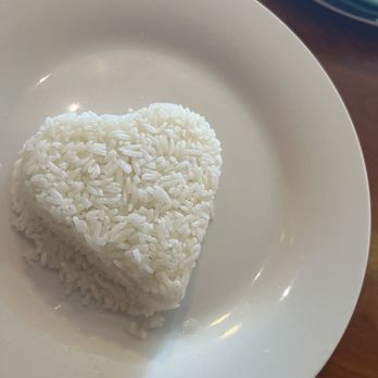 Thai Tip - Heart shaped rice - nice touch - A serving of white rice shaped like a heart is presented on a plain white plate. The rice is neatly molded, and the plate is set on a wooden surface. A blue bowl is partially visible in the upper right corner of the image, indicating a dining setting. - Thai Near Me - Albuquerque, New Mexico
