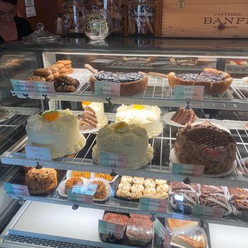 Patty B's - A glass bakery display case contains multiple shelves with a variety of baked goods, including cheesecakes, chocolate chip cookies, ricotta cookies, and other desserts. The case is situated in a bakery or cafe environment, with a person visible in the background and labels identifying the items. The display emphasizes homemade-style cakes and cookies, with some decorated with frosting and toppings. - Italian Near Me - Dover, New Hampshire
