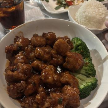 All Seasons Table - General Gaus Chicken - A white bowl contains sweet and sour chicken coated in a glossy sauce, with pieces of broccoli on the side. The setting includes a glass of soda with ice, a plate of salad with greens and cherry tomatoes, and a bowl of white rice. The table appears to be set for a meal at a restaurant. - Chinese Near Me - Malden, Massachusetts