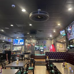 Interior - The image shows the interior of a casual sports bar with wooden flooring, tables, and chairs arranged for patrons. Several flat-screen TVs are mounted on the walls, displaying sports events and other programs. The ceiling is dark with recessed lighting and a large ceiling fan. In the background, gaming machines and a bar area are visible, along with framed pictures and decorations on the walls. The atmosphere appears relaxed and suitable for watching sports and socializing.