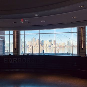 Hyatt Regency Boston Harbor - View from hotel lobby - The image shows an indoor space with a curved window wall providing a view of a city skyline across a river. The interior is dimly lit, with some ceiling lights visible. Outside, a person is walking along the waterfront, and city buildings are visible in the background. The window has string lights hanging across it, and the space appears to be part of a restaurant or lounge area. - Hotels Near Me - Boston, Massachusetts