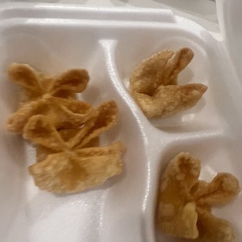 Rice Cafe - Fried Wontons (it comes with 6) - Three fried wontons are placed in a foam takeout container with three separate compartments. The wontons are golden-brown and crispy, typical of fried appetizer dishes. The container appears to be on a flat surface, possibly a table or counter, with a white background. The image focuses on the food, which is part of a meal from a restaurant offering Asian cuisine. - Sushi Bars Near Me - Madison, Mississippi