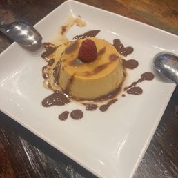 Flan - A caramel flan dessert topped with a single raspberry is presented on a white square plate. The flan is drizzled with chocolate sauce, which is also spread around the plate. Two metal spoons are placed on opposite sides of the plate, and the dessert is set on a dark wooden table. The presentation emphasizes the contrast between the flan's caramel color and the raspberry, with decorative chocolate accents.