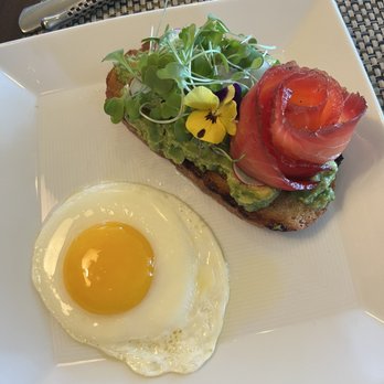 JORY - Avocado toast - A square white plate holds avocado toast topped with microgreens, a yellow edible flower, and a rolled slice of smoked salmon. A fried egg with a runny yolk is placed beside the toast. The dish is set on a dark table with a woven placemat, and a knife and fork are positioned above the plate. - New American Near Me - Newberg, Oregon