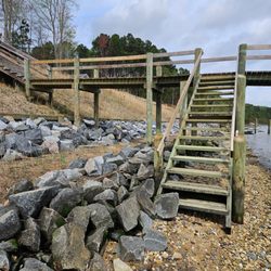 Wooden stairs descend from an elevated boardwalk to a rocky shoreline. Treated lumber posts support the structure, while larg...