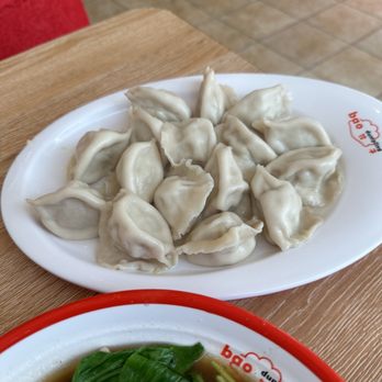 Bao Dumpling - Pork Cabbage & Pork 16pic - A white plate with steamed dumplings is placed on a wooden table at Bao Dumpling, a Chinese restaurant. Part of a bowl with broth and greens is visible. - Chinese Near Me - West Lafayette, Indiana