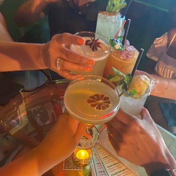 Bar Nancy - A group of people is holding cocktails with lemon slices and garnishes, celebrating at a bar with a dark, ambient atmosphere. - Cocktail Bars Near Me - Miami, Florida