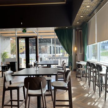 Placebo - tables and chairs in a restaurant - The image shows an interior space of a restaurant or bar with high tables and chairs arranged near large windows that allow natural light to fill the room. The decor includes dark walls, green curtains, and a patterned ceiling panel. The floor is wooden, and outside the windows, a patio and building structures are visible. The space appears modern and clean, suitable for casual dining or drinks. - Bars Near Me - Cincinnati, Ohio