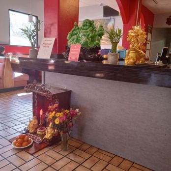 Việt kitchen - A little of the interior view - A restaurant interior features a counter with decorative statues, including a green laughing Buddha and a gold figure, along with potted plants. On the floor, there is a vase of flowers, a plate of oranges, and small statues with candles. The background includes a window, seating area, and a red wall with an archway. - Vietnamese Near Me - Albuquerque, New Mexico