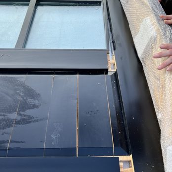 Rustica - The image shows a flooring installation process with black tiles laid on a wooden subfloor. A metal frame supports the tiles, and some tiles are already set, while others are positioned for installation. Bubble wrap covers the side area, likely for protection during the process. Hands are visible on the right side, indicating ongoing work. - Home Decor Near Me - Springville, Utah