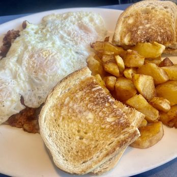 Airway - Hubby's brunch - A white oval plate contains two fried eggs with runny yolks, two slices of toasted bread, and a portion of fried potato chunks. The eggs are positioned on the left side of the plate, with the toast slices placed at the top and bottom, and the potato chunks filling the center and right side. The meal appears to be served in a casual restaurant or diner setting, with a dark table surface and a cloth napkin partially visible in the background. - Breakfast & Brunch Near Me - Windsor Locks, Connecticut