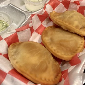 Manny's Cabin - (Pasteles) Mixed Mini wheat empanadas. Cheese, Chicken and Beef - Three empanadas with golden, flaky crusts are arranged on red and white checkered paper inside a food basket. Small plastic containers of sauce are visible in the background, along with a metal tray and a cup of sauce. The setting suggests a casual dining or takeout environment, typical for Venezuelan eateries offering traditional snacks like empanadas. - Venezuelan Near Me - Florence, Kentucky