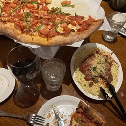 Pizza & pasta - The image shows a wooden table with a large pizza featuring pepperoni and green peppers, with a slice removed. There is a plate of spaghetti topped with meat sauce and garnished with herbs, along with a glass of red wine and a glass of water. Part of a person's arm and clothing are visible in the background, indicating a dining setting.