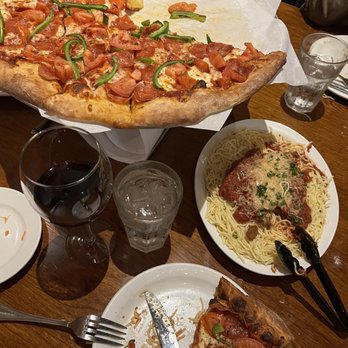 Franky's - Pizza & pasta - The image shows a wooden table with a large pizza featuring pepperoni and green peppers, with a slice removed. There is a plate of spaghetti topped with meat sauce and garnished with herbs, along with a glass of red wine and a glass of water. Part of a person's arm and clothing are visible in the background, indicating a dining setting. - Italian Near Me - Chico, California