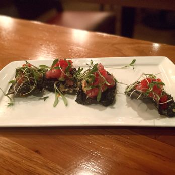 Scratch - Ahi Poke - Three pieces of dark-colored food topped with microgreens and diced tomatoes are arranged on a white rectangular plate, set on a wooden table. - New American Near Me - Mountain View, California