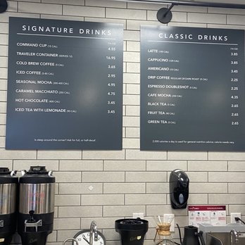 Command Coffee - Menu - Two large menu boards are mounted on a tiled wall in a coffee shop, listing various signature and classic drinks with prices and calorie information. The signature drinks include options like command cup, traveler container, and seasonal mocha, while the classic drinks feature items such as latte, cappuccino, and black tea. Below the boards, a coffee station with brewing equipment, a sink, and various accessories is visible. The setting appears clean and organized, with a neutral color scheme and functional layout. - Coffee & Tea Near Me - Indianapolis, Indiana