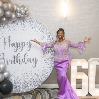 MNPhotoStudios - A woman wearing a purple gown with lace details poses with arms outstretched in front of a birthday celebration backdrop. The backdrop features a large circular sign with 'Happy Birthday' written on it, surrounded by silver and black balloons. To her right, a large illuminated '60' sign is displayed on the floor. The setting appears to be indoors with neutral-colored walls and soft lighting. - Session Photography Near Me - Chicago, Illinois