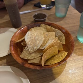 Los Arcos - Chips & Salsa - A wooden bowl containing tortilla chips is placed on a restaurant table with drinks and condiments in the background. - Tacos Near Me - Chicago, Illinois