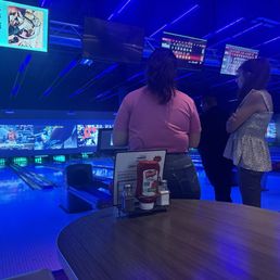 Three women are gathered near the bowling lanes in a bowling alley with multiple screens displaying scores and advertisements.