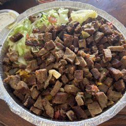 Lamb Platter - A large aluminum tray holds chopped cooked meat, possibly beef or lamb, with visible seasoning and small vegetable pieces. Lettuce and other vegetables are placed on one side of the tray. The tray is on a wooden table, with a small container of sauce or dressing nearby, indicating a casual dining setting.