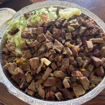 Halal Gyro Master - Lamb Platter - A large aluminum tray holds chopped cooked meat, possibly beef or lamb, with visible seasoning and small vegetable pieces. Lettuce and other vegetables are placed on one side of the tray. The tray is on a wooden table, with a small container of sauce or dressing nearby, indicating a casual dining setting. - Halal Near Me - River Vale, New Jersey