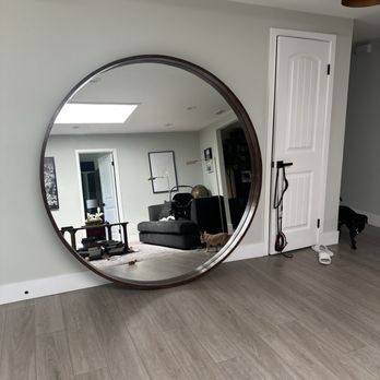 OC Fixit - Previously unhangable mirror - A large, round mirror is positioned on the floor, reflecting a living room with a gray sofa, artwork, and a skylight. The room has light-colored walls and wooden flooring. - Furniture Assembly Near Me