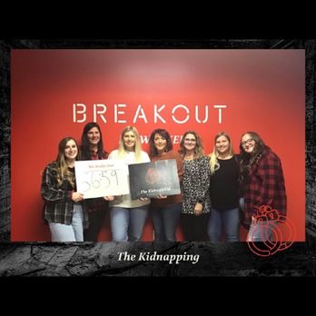 Breakout Games - Six women stand together in front of a red wall with the word 'BREAKOUT' displayed prominently. They are holding signs, one with the time '5:59' and another with the logo 'The Kidnapping.' The setting appears to be an escape room venue, and the photo captures a group celebrating or commemorating their experience. The image is framed with a black border and a small graphic of a bomb in the lower right corner. - Escape Games Near Me - Brookfield, Wisconsin