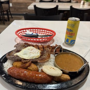 Aroma Cafe - Bandeja Paisa was so good. - The image shows a Colombian-style meal on a black tray, featuring white rice, a fried egg on grilled beef, slices of sausage, fried plantains, and a small bowl of beans. A can of soda is placed behind the tray, and the setting appears to be a casual restaurant with white tables and black chairs, decorated with small potted plants. - Colombian Near Me - Springfield, Virginia