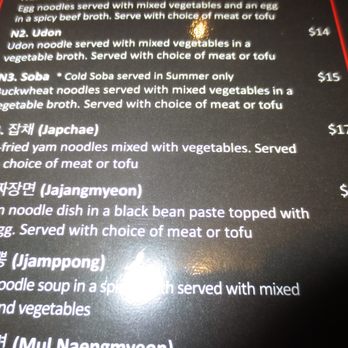 Kimchi - A photograph of a Korean restaurant menu featuring a list of noodle dishes including Udon, Soba, Japchae, Jajangmyeon, and Jjamppong. Each dish has a description of ingredients and preparation style, along with prices ranging from $14 to $17. The menu is printed on a dark background with white text and red borders. - Korean Near Me - Ithaca, New York