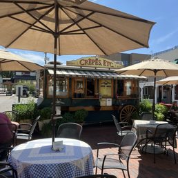 Michel's Vorner - A food truck with a sign reading 'Crepes, Etc.' is situated in an outdoor patio area. The space includes tables with checkered and patterned tablecloths, shaded by large umbrellas. Potted plants decorate the area, and there are a few patrons seated at the tables. The sky is clear and blue, indicating a sunny day.