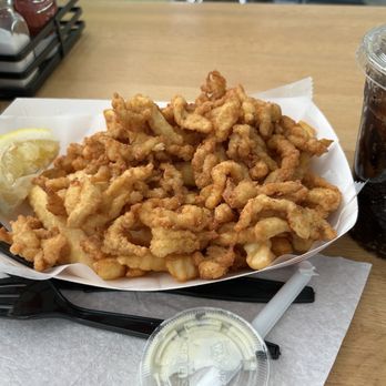 Murphy's Eats and Treats - Clam Strip Dinner 15.95 - A serving of fried calamari, consisting of battered and deep-fried squid pieces, is presented in a paper-lined basket on a wooden table. A lemon wedge is placed on the side for flavor. A plastic cup filled with a dark soda and a straw is visible to the right. The setting appears to be a casual dining restaurant with condiments in the background. - Burgers Near Me - Framingham, Massachusetts