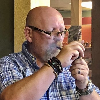 Cigar Inn - A man with a bald head, glasses with a tortoiseshell pattern, and a beard is lighting a cigar using a lighter. He is wearing a plaid shirt and multiple bracelets on his wrist. The background features a wooden structure and a decorative sculpture, indicating an indoor setting such as a tobacco shop or lounge. - Tobacco Shops Near Me - Phoenix, Arizona