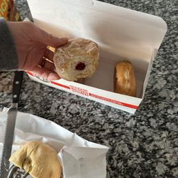 Kolache - A hand, wearing a gray sleeve, holds a partially eaten jelly-filled donut with powdered sugar over a white box containing two other donuts, one of which is a glazed donut. The donuts are placed on a dark granite countertop. Nearby, there is a serrated knife on a paper bag, a small bottle of chocolate syrup, and a box of Froot Loops cereal. The scene suggests a casual snack or breakfast setting.