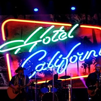 Farnsworth Hall - Eagles Tribute. - A musical band is playing on stage with a large neon sign that says 'Hotel California' behind them, illuminated with colorful lights. - Music Venues Near Me - Mesa, Arizona