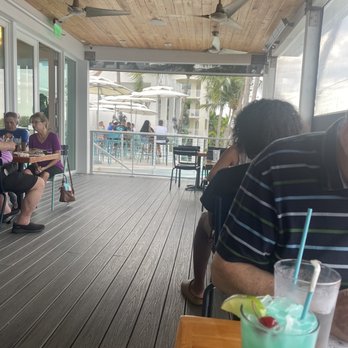 TideHouse - Outside with a view of the patio with umbrellas - The image shows an indoor dining area with a wooden ceiling and ceiling fans, adjacent to a large window or open wall that overlooks an outdoor patio with umbrellas and palm trees. Patrons are seated at tables, some enjoying meals and drinks. In the foreground, a person is partially visible, and a colorful cocktail with a cherry and lemon wedge is on the table. The setting appears to be a waterfront restaurant with a relaxed atmosphere. - Seafood Near Me - Stuart, Florida