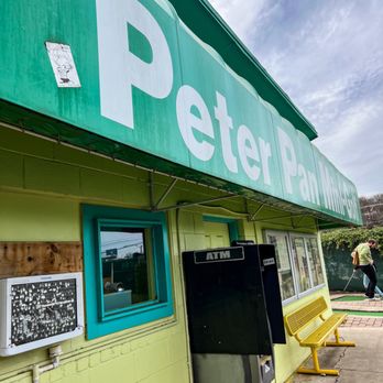 Peter Pan Mini-Golf - The photo shows the front exterior of Peter Pan Mini-Golf in Austin, TX, featuring a green building with a large sign reading 'Peter Pan Mini-Golf.' There is an ATM machine and a yellow bench outside. A person is seen walking in the background near the sidewalk. The sky is partly cloudy, and the scene captures a casual outdoor setting near the mini-golf venue. - Mini Golf Near Me - Austin, Texas