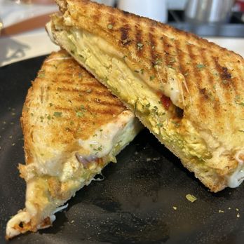Llamas’ Corner - Llama Sunrise Breakfast Panini - A grilled sandwich with golden-brown toasted bread, filled with melted cheese, avocado slices, and herbs. The sandwich is cut in half, revealing the creamy filling inside, and is served on a black plate. The background shows a blurred cafe setting with various items. - Themed Cafes Near Me - Baltimore, Maryland
