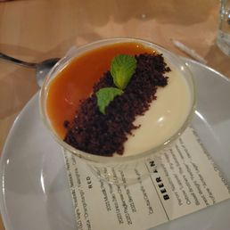 Panna cotta - A panna cotta dessert in a clear glass cup features a creamy white base topped with a layer of caramel sauce. It is garnished with a line of chocolate crumbs and two small mint leaves. The dessert is served on a white plate with a printed paper underneath, on a wooden table. A spoon and a glass of water are visible nearby, and a person's arm with a watch is in the background.