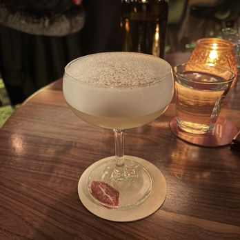 Bisous - A cocktail with foam on top and a garnish at the base is placed on a wooden table in a bar setting, with a water glass and candle nearby. - Cocktail Bars Near Me - Chicago, Illinois