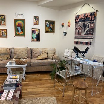 Luna and Luz - A cozy interior space featuring a beige sofa, various colorful artworks on the wall, and display furniture including a clear acrylic desk and a small wooden stool. - Jewelry Near Me - Albuquerque, New Mexico