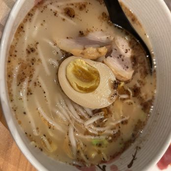 Noku - Very bland tasteless ramen - A close-up of a bowl of ramen containing a halved soft-boiled egg, slices of meat, noodles, and a creamy broth with black pepper and small bubbles. A black spoon rests inside the bowl, which is placed on a wooden surface. - Korean Near Me - Reston, Virginia