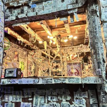 Hideout Saloon - Amazing decor - The image shows the interior of a bar or club with walls, ceiling, and fixtures densely decorated with dollar bills, graffiti, and drawings. The space features wooden beams and a ceiling with lighting fixtures. The decor includes various handwritten messages, drawings, and stickers, giving it a chaotic, artistic appearance. The bar area is visible with some equipment and signage, emphasizing a casual, unconventional ambiance. - Cocktail Bars Near Me - Mariposa, California