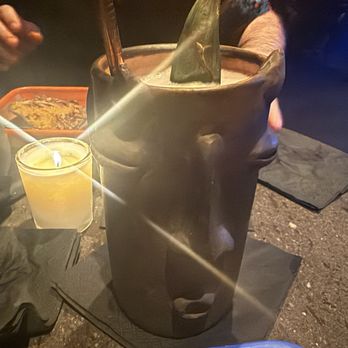 La Mezca - A large black ceramic mug with a face design, including eyes, nose, and mouth, is on a table. The mug contains a beverage with a straw and a lime wedge. Nearby, there is a lit candle in a glass holder, a small bowl, and a food container. The setting appears to be a dimly lit restaurant or bar with a dark table surface and black napkins. - Cocktail Bars Near Me - Austin, Texas