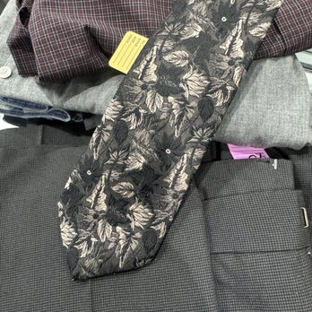 Second Chance Thrift - The image shows a pair of fabric pants with a black and beige leaf pattern, placed on a surface among other clothing items, including a gray garment and a checkered fabric. The pants have a button and zipper closure, and the pattern features detailed leaf motifs. The surrounding clothing items are folded and include various textures and colors, indicating a clothing display or selection. - Thrift Stores Near Me - Glendora, California