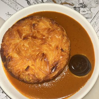 Eyval - Tahdig - A baked potato topped with shredded cheese is served on a plate with brown gravy and a small container of sauce, set on a table with printed paper. - Persian/Iranian Near Me - Brooklyn, New York
