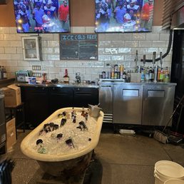 Bar - Bar interior featuring two mounted TVs displaying a football game, a chalkboard menu, and a bathtub filled with bottles of beer on ice.
