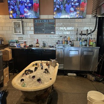 Firewater BBQ - Bar - Bar interior featuring two mounted TVs displaying a football game, a chalkboard menu, and a bathtub filled with bottles of beer on ice. - Barbeque Near Me - Elmhurst, Illinois